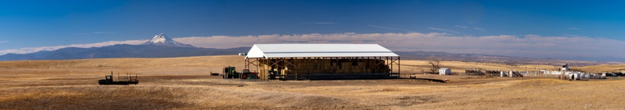 A panorama image of a hay storage barn and farm equipment and rodeo grounds and  on a farm near Dufur Oregon with Mt Hood in the background.