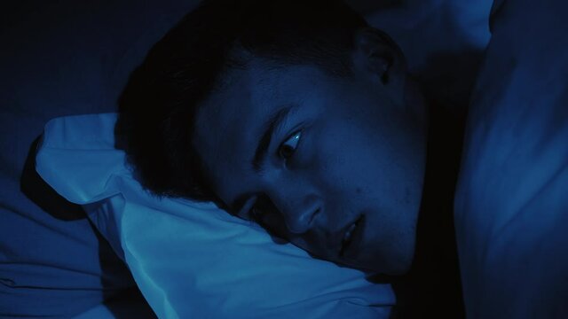 Scary nightmare. Sleep disorder. Restless night. Disturbed frightened man woke up opened eyes lying in bed late at home in dark blue light.