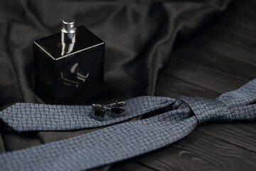 A bottle of mens cologne and cufflinks with blue tie lie on a black luxury fabric background on a...