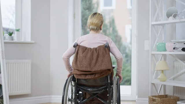 Back view of disabled young woman rolling wheelchair to glass door at home. Handicapped Caucasian paraplegic lady indoors on spring or summer day. Hopelessness and disability.