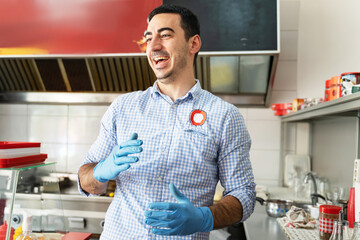 Young adult caucasian man entrepreneur standing at his fast food store in day smiling front view Chef in the kitchen having fun at work wearing protective medical gloves to prevent disease real people