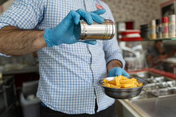 Close up on hands midsection of unknown man entrepreneur with blue rubber protective glove salting potato chips french fries in metal plate at the fast food restaurant in day serving