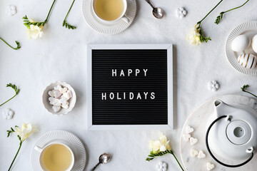 Text Happy Holidays on letter board with spring freesia flowers and tea cup on table. Black...