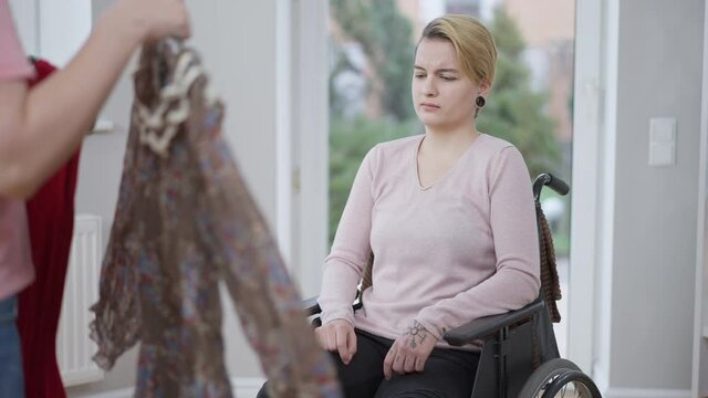 Portrait of young tattooed paralyzed woman sitting in wheelchair as unrecognizable blurred friend showing new dresses. Sad depressed Caucasian disabled invalid choosing outfit at home.