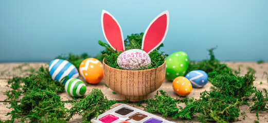 Bright colorful Easter eggs and rabbit ears on a blue and wooden background with green moss