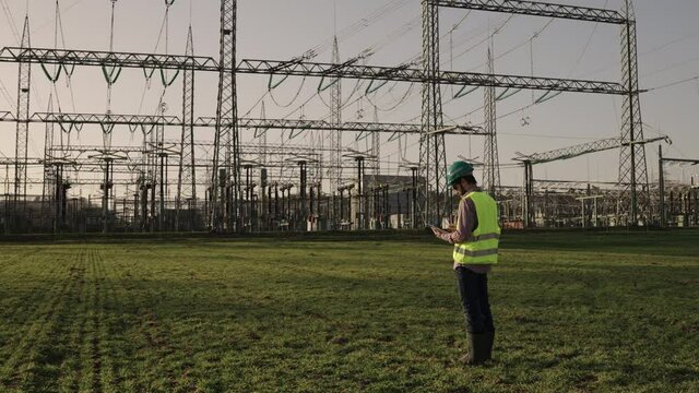 Electrical engineer wearing a helmet and safety vest working with tablet near high voltage electrical lines power station during sunset shot in 4k super slow motion
