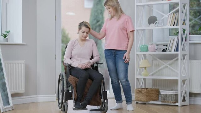 Wide shot of empathic woman caressing hair and hugging disabled friend in wheelchair. Young Caucasian lady taking care supporting paralyzed invalid. Slow motion.