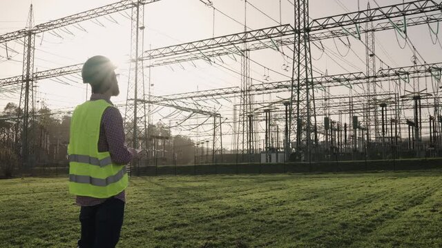 Electrical engineer wearing a helmet and safety vest working with tablet near high voltage electrical lines power station during sunset shot in 4k super slow motion