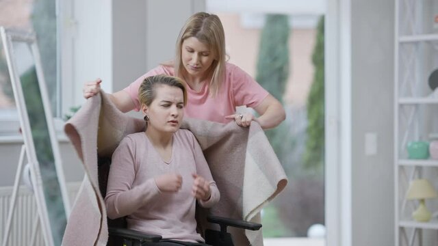 Angry stressed paralyzed woman taking off blanket as friend taking care of disabled person. Portrait of frustrated Caucasian invalid refusing help. Disability problems and lifestyle.
