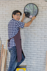 asian male adult wearing clear safety glasses and blue plaid shirt holding and adjusting black round metal clock with white numbers and hands on white brick wall and stand smiling happily