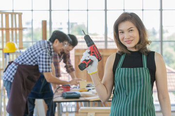 asian adult female worker wearing black shirt with green stripe apron stand smiling and holding red electric screwdriver when her male colleagues wearing plaid shirt discussing behind at wooden table 
