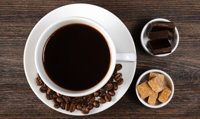White porcelain cup of coffee, saucer with coffee beans, bowls with cane brown sugar and dark chocolate on dark wooden background