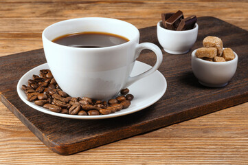 White porcelain cup of coffee, coffee beans, bowls with cane brown sugar and dark chocolate on dark wooden background. Selective focus