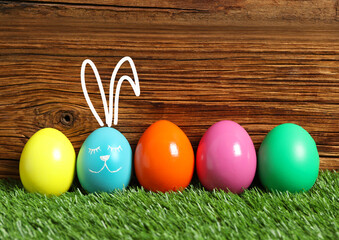 One egg with drawn face and ears as Easter bunny among others on green grass against wooden background