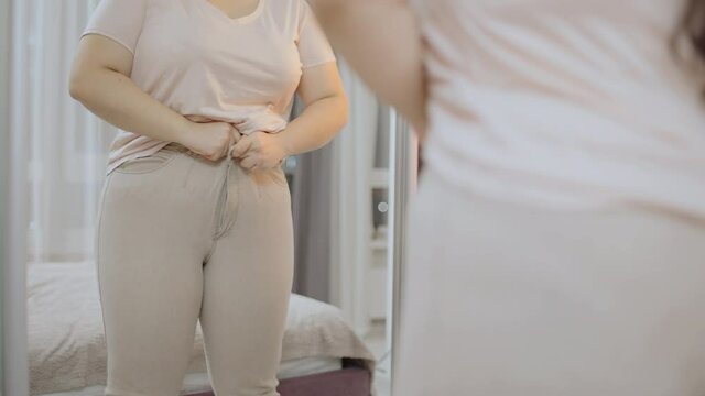 Obese female trying to zip up tight trousers in front of mirror, plus size