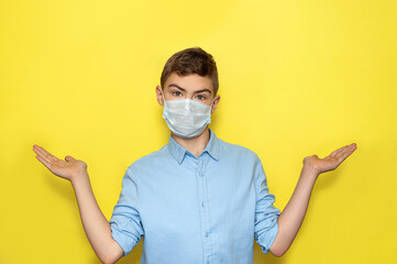 The boy was a teenager in a medical mask and a blue shirt on a yellow background spread his hands in different directions. The boy points to two objects. Surprise.