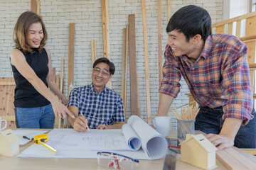 Middle aged man wearing blue plaid shirt holding pencil explaining and discussing blueprint document to young male and female colleagues on the working table in construction room