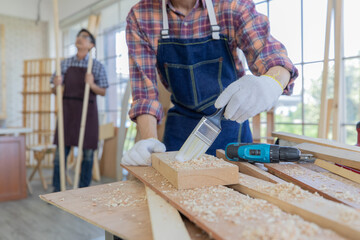 Young asian male worker wearing plaid shirt and blue jean apron with white gloves holding saw cutting wood stick on the table full of building tools and equipments while his colleague stand holding 