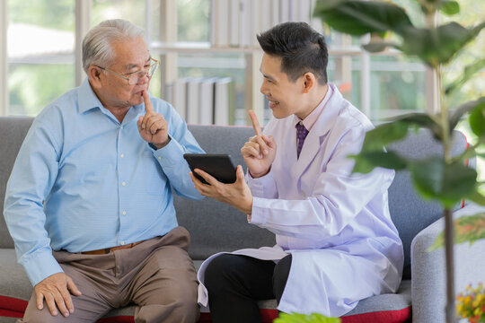 A young asian male doctor wearing white lab coat holding tablet sitting on sofa talking and discussing with old fat patient sitting next to him when both people hold index finger 