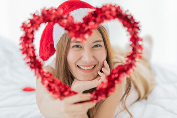 Close up of a young asian long brown hair female face wearing red and white santa hat lay down on white sheet bed through heart shape shiny ribbon object