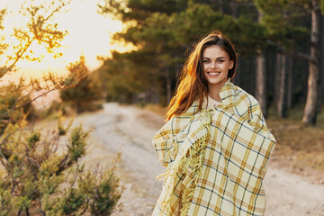 A woman with a warm blanket on her shoulders smiles near the green bushes in nature in the forest