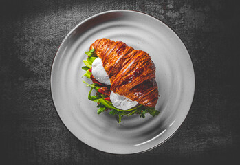Croissant sandwich with poached egg, tomato and avocado on plate on Dark grey black slate background