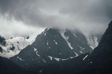 Fototapeta na wymiar Dark atmospheric mountain landscape with glacier on black rocks in lead gray cloudy sky. Snowy mountains in low clouds in rainy weather. Gloomy landscape with black rocky mountains with snow in fog.
