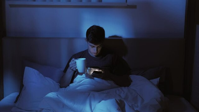 Sleepless night. Internet insomnia. Late online. Relaxed guy enjoying reading feed using mobile phone drinking sedative herbal tea alone in bed at home in dark blue light with defocused effect.