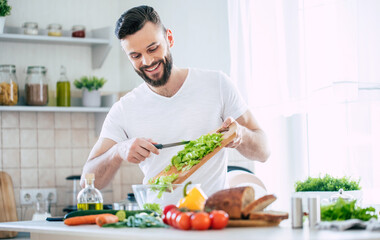 Handsome happy bearded man is preparing wonderful fresh vegan salad in the kitchen at home - obrazy, fototapety, plakaty
