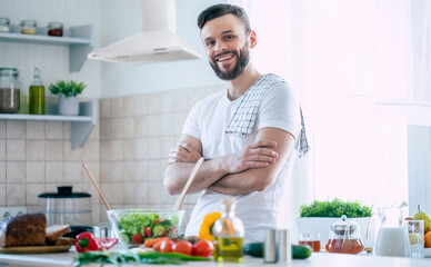 Handsome muscular young bearded smiling man in the kitchen at home is looking on the camera with cross hands