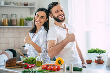 Excited smiling young couple in love making a super healthy vegan salad with many vegetables in the kitchen and showing thumbs up