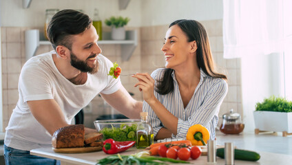 Excited smiling young couple in love making a super healthy vegan salad with many vegetables in the kitchen and man testing it from a girl's hands