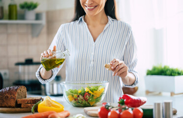 Close up photo of a smiling young woman makes a fresh vegan salad while she uses olive oil for it.