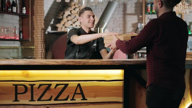 Smiling pizzaiolo in black uniform giving three pizza boxes to male client at restaurant. Happy man taking tasty italian food from modern pizzeria.