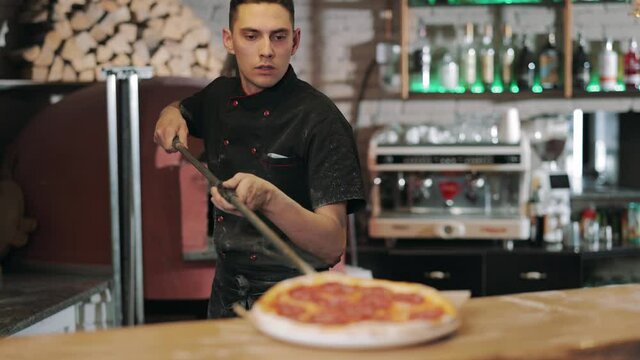 Competent Chef Cook In Black Uniform Taking Out Of Stove Hot Baked Pizza Using Long Shovel. Pizza Maker Serving Tasty Italian Meal On Round Plate At Restaurant.