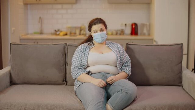 Young obese woman in mask looking at belly fat, quarantine weight gain, body