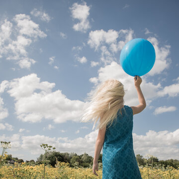 rear view of blonde girl in blue dress with a blue balloon  in summer or spring under a blue sky with clouds