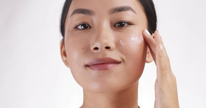 Close up portrait of young asian lady applying cream on cheek, white studio background, slow motion