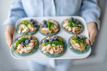 Woman in blue shirt holding a set of american pancakes on a plate with seaweed, cheese, blueberry and hummus. Homemade tasty snack.