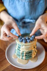 Woman in blue apron decorate blue american pancakes with blueberry poured with caramel souse on wood table. Homemade tasty food. Celebration of Shrovetide.