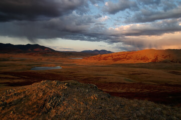 Dramatic sunset highland steppe valley with lakes with dry yellow grass on the background of rocky mountains under storm sky