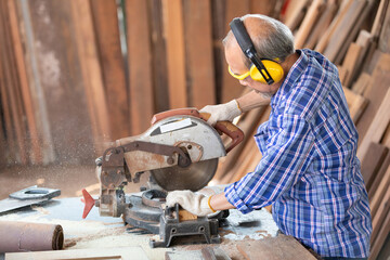 senior asian man carpenter wearing glasses and headphone, using electric circular saw for cutting wooden boards, on a piece of wood in workshop