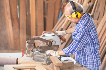 senior asian man carpenter wearing glasses and headphone, using electric circular saw for cutting wooden boards, on a piece of wood in workshop