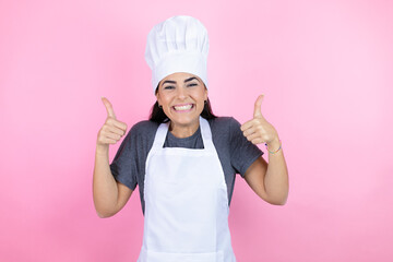 Young hispanic woman wearing baker uniform over pink background success sign doing positive gesture with hand, thumbs up smiling and happy. cheerful expression and winner gesture.