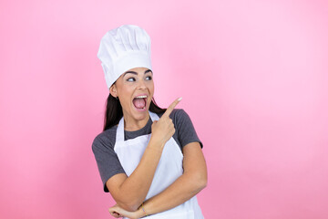 Young hispanic woman wearing baker uniform over pink background amazed and pointing with hand and finger to the side