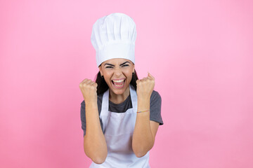 Young hispanic woman wearing baker uniform over pink background very happy and excited making winner gesture with raised arms, smiling and screaming for success.