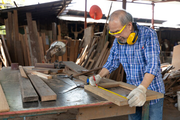 senior asian man carpenter wearing glasses and headphone, measuring wood with tape measure in workshop