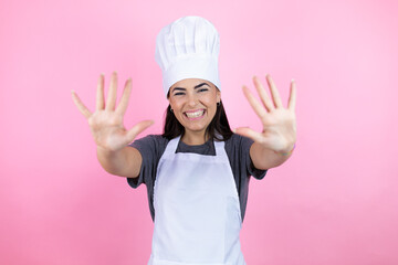 Young hispanic woman wearing baker uniform over pink background showing and pointing up with fingers number ten while smiling confident and happy