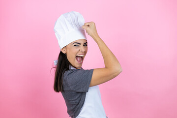 Young hispanic woman wearing baker uniform over pink background feeling happy, satisfied and powerful, flexing fit and muscular biceps, looking strong after the gym