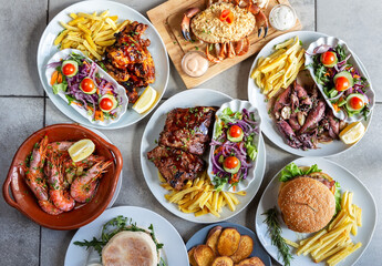Diverse plates of mediterranean and western food ranging from shrimp, crab, squid, ribs, a hamburger, lots of salad and fries.